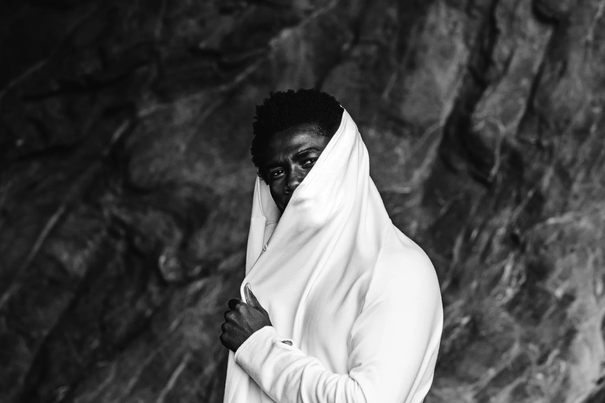 Copala model draping a white garment across his face against dark wet rock, dramatic black-and-white menswear editorial by Chauncey Scott, Leo Carrillo State Beach, Malibu